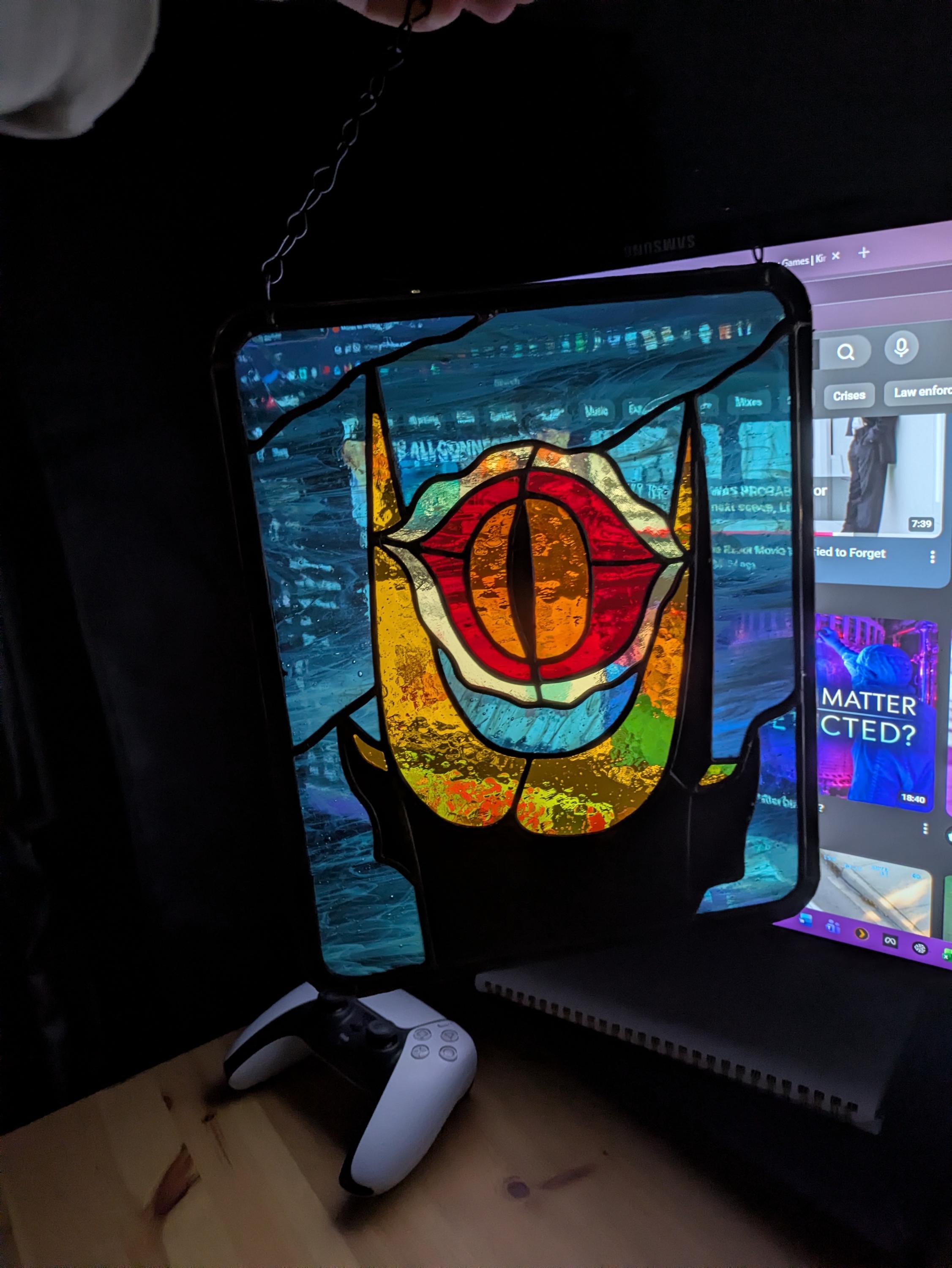 It Watches stained-glass panel with a red and amber eye, yellow hand and blue background, lit by a screen